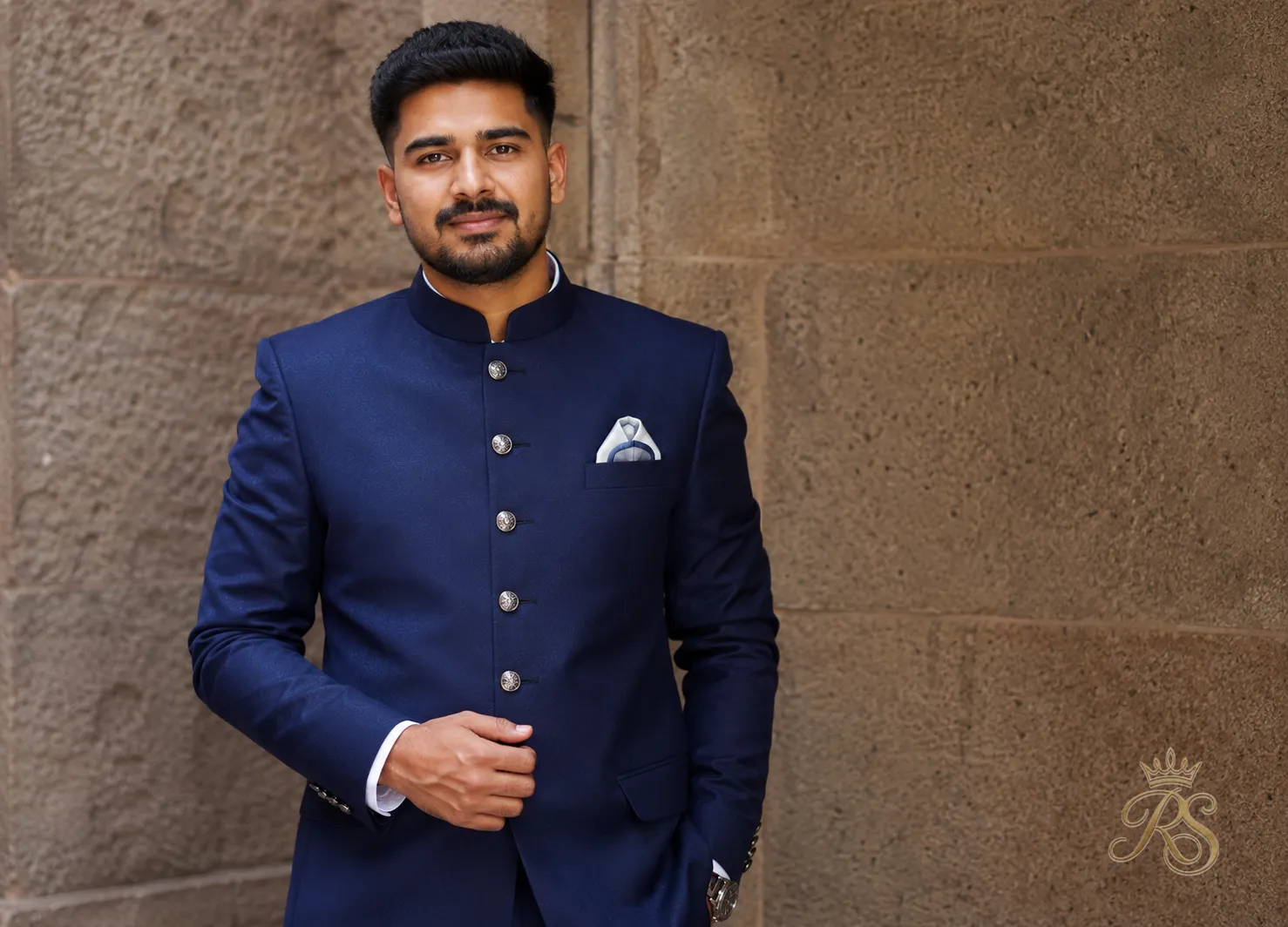 Groom in a navy bandhgala suit with a pocket square standing against a stone wall