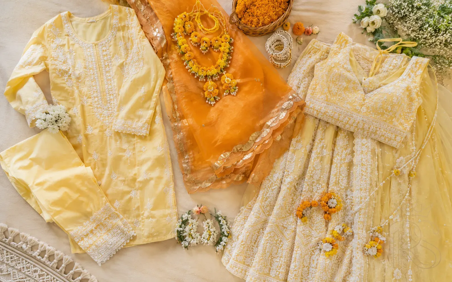 Flat lay of yellow and orange Haldi ceremony outfits including chikankari kurta set, organza saree and lemon lehenga with floral accessories