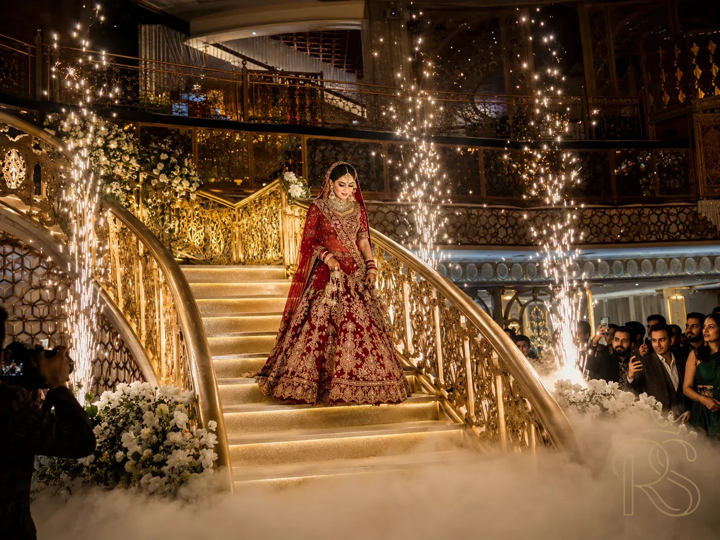 Dramatic Indian bridal entry down a grand staircase — bride in deep red and gold lehenga descending gilded stairs surrounded by cold pyrotechnic sparklers, fog machine effect, wedding guests looking on, professional wedding photography, cinematic lighting, wide angle shot