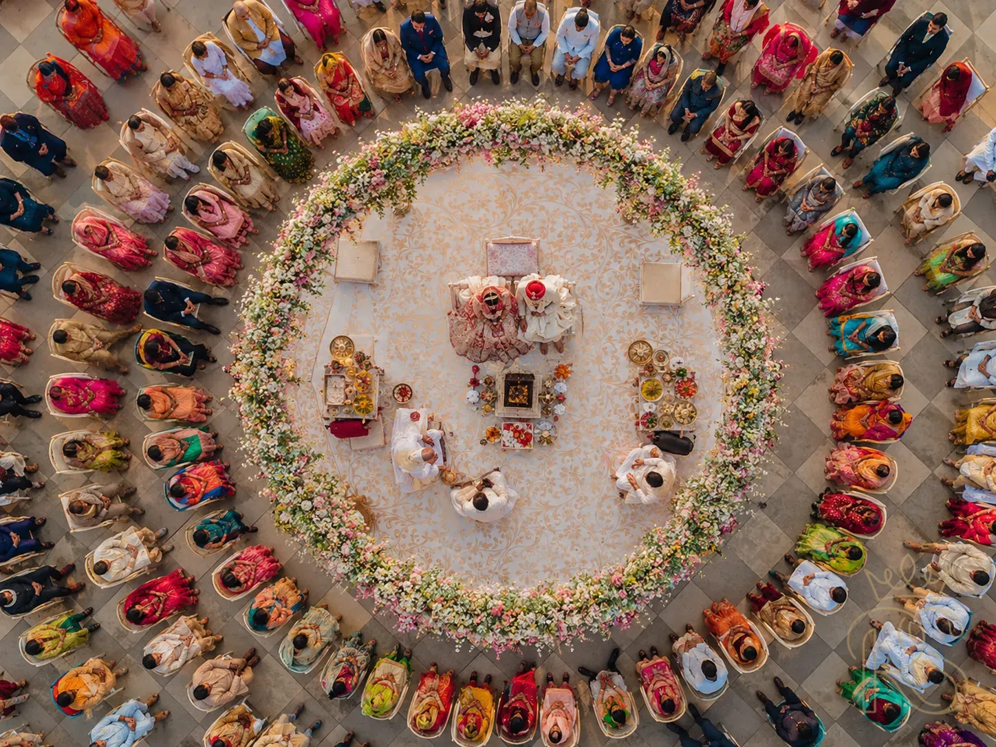 Aerial drone photograph of an Indian wedding ceremony from directly above — circular mandap with flowers, bride and groom at centre, guests seated in concentric rows around them, top-down bird's eye view, vivid colours, editorial wedding photography
