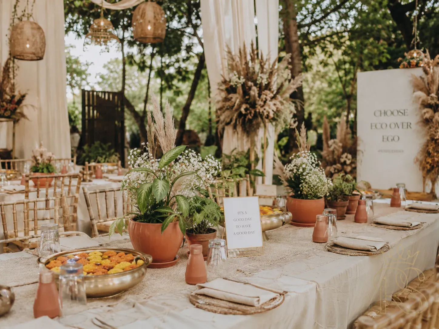 Eco-friendly Indian wedding setup with potted plants as table centrepieces, natural cotton fabric draping, terracotta pots, dried flowers and pampas grass, brass urlis with floating marigolds, bamboo furniture, outdoor garden setting, warm natural light, editorial sustainability-focused photography