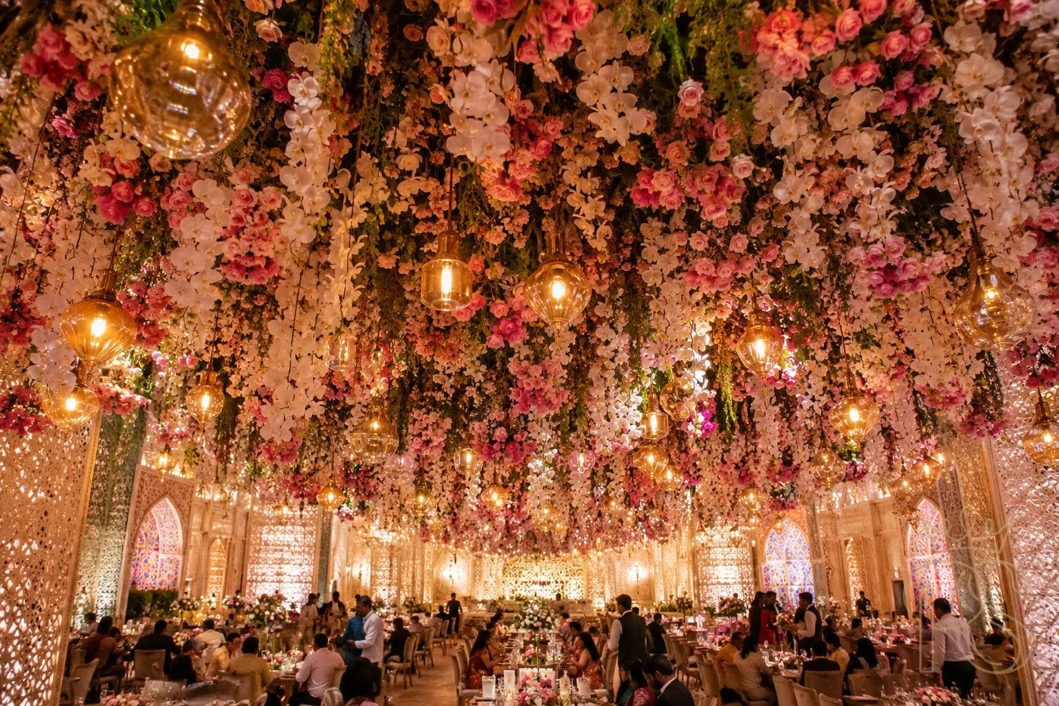 Breathtaking Indian wedding hall with a full floral ceiling installation — thousands of white orchids, pink garden roses, and trailing greenery suspended overhead creating a floral canopy, warm amber pendant lights interspersed, guests dining below
