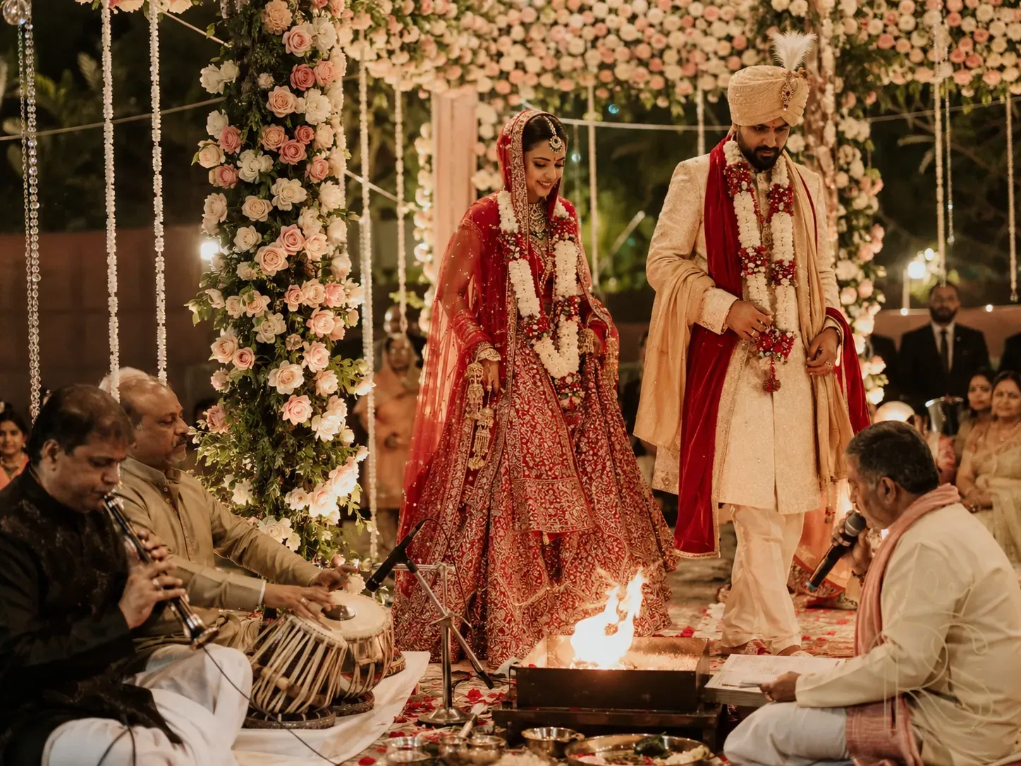 Emotional Indian wedding pheras ceremony with live musical performance — musicians playing shehnai and tabla beside the havan kund, bride and groom walking around the sacred fire, priest chanting, intimate emotional atmosphere, soft warm lighting, candid documentary photography