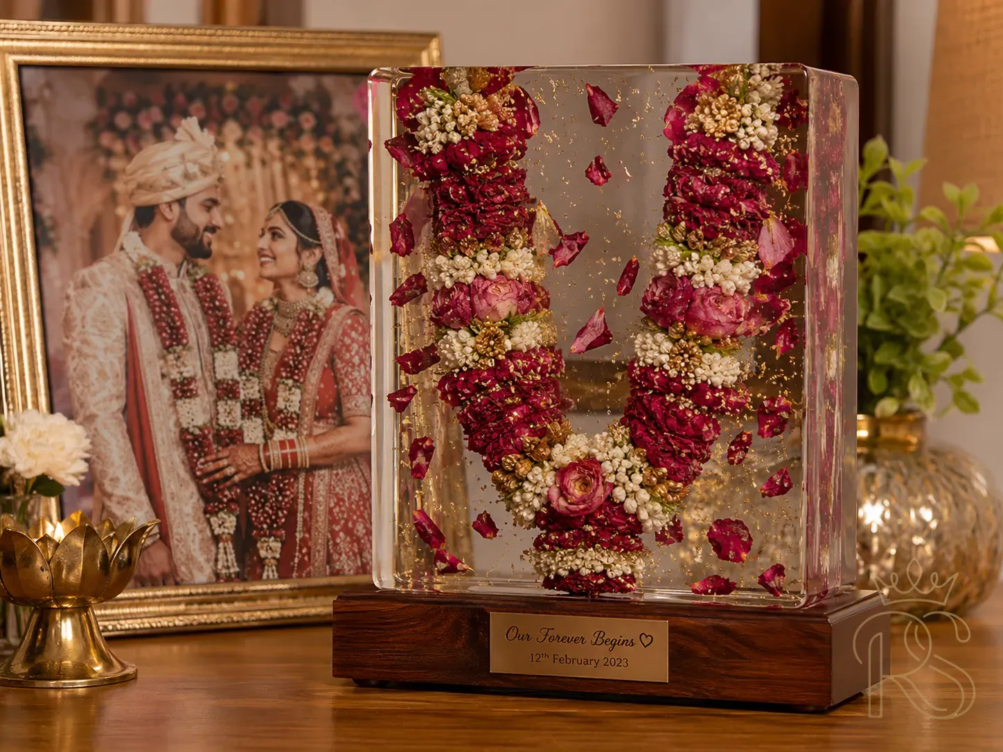 Beautiful Indian wedding varmala (flower garland) preserved in clear resin — embedded in a rectangular resin block with rose petals visible inside, placed on a wooden display base, styled with the couple's wedding photograph behind it, warm home decor photography