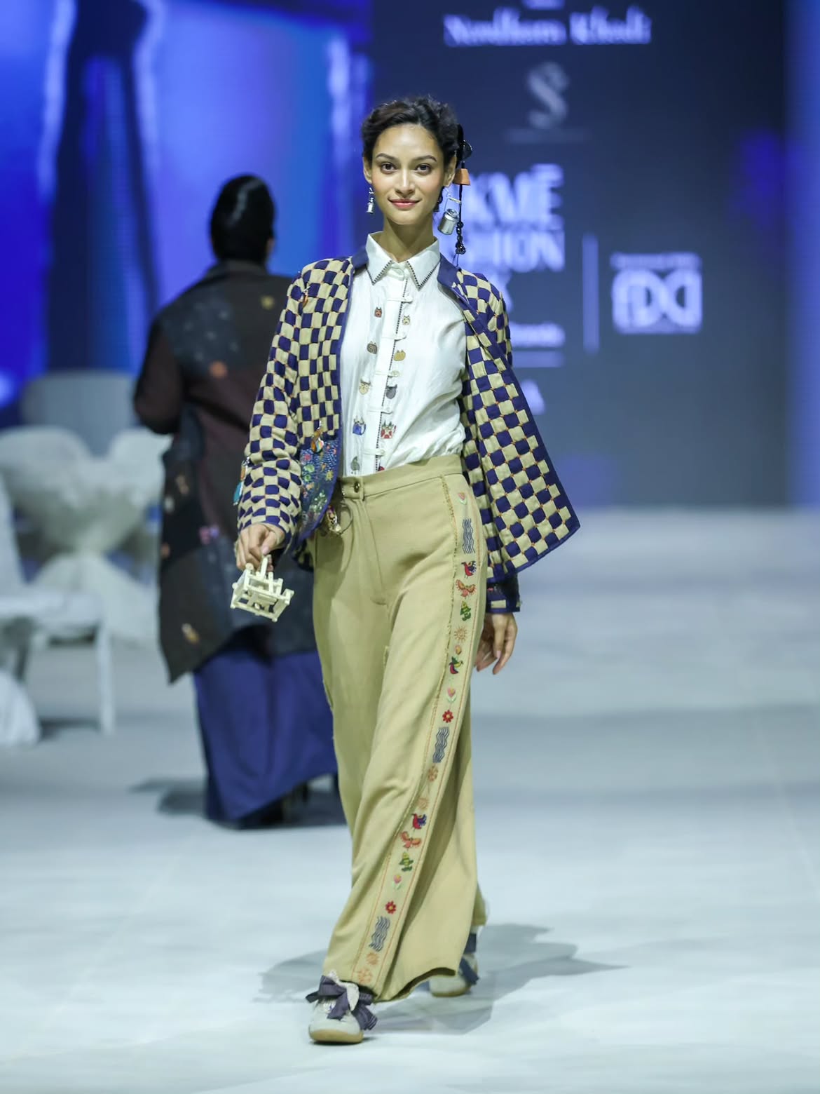 Model on the runway in a checkerboard jacket with embroidered charms, over a white shirt and khaki trousers