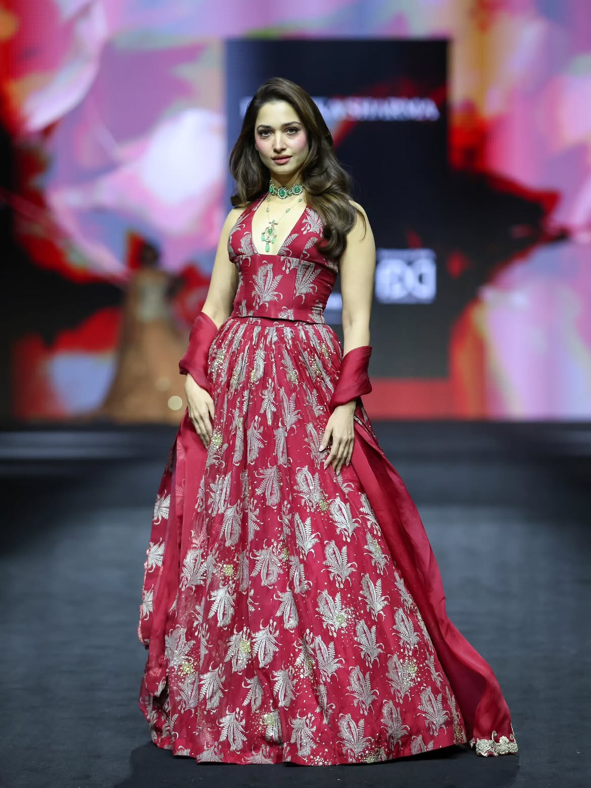 Tamannaah Bhatia walks the runway for Bhumika Sharma in a deep red embroidered ball gown with emerald jewelry