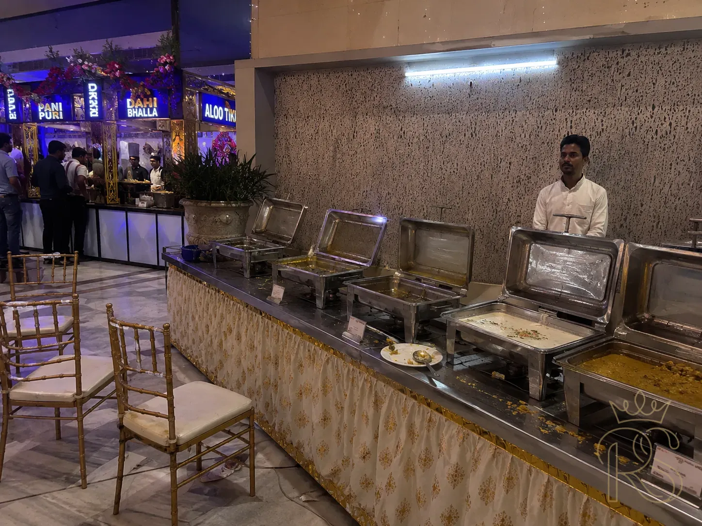 Long abandoned Indian wedding buffet station with barely touched food — stainless steel chafing dishes with congealing food, empty chairs, one lonely attendant standing by, fluorescent overhead light, contrast to the lively chaat stations nearby — illustrative photography, slightly desaturated tones to convey the flat mood, realistic wedding catering setting