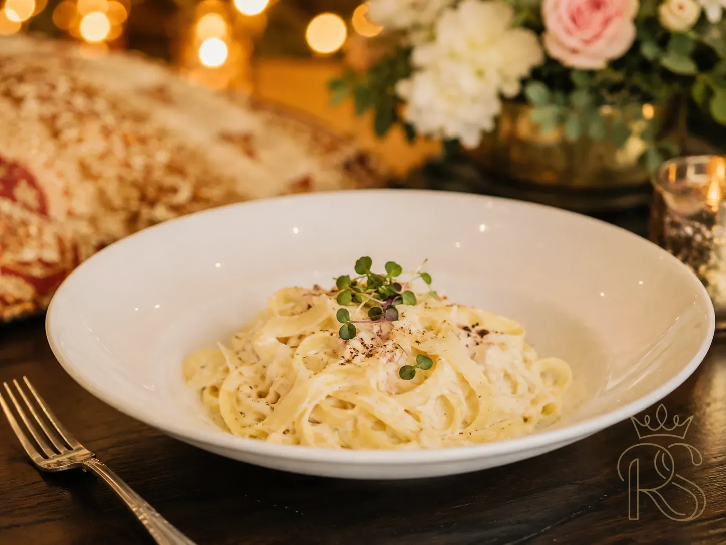 Elegant plated pasta bowl at a luxury Indian wedding — creamy fettuccine alfredo garnished with microgreens, cracked black pepper, shaved parmesan, served in a white ceramic bowl on a dark wood surface with a small fork, blurred fairy lights in background, editorial food photography, natural warm light, clean minimalist plating