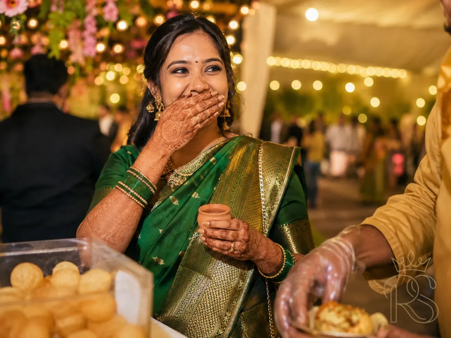 Candid Indian wedding photograph of a woman in a rich green silk saree laughing with delight while eating pani puri at a live wedding food counter, hand covering mouth, small clay kulhad in other hand, blurred fairy light bokeh behind her, warm golden evening light, photojournalism style, joyful expression, candid wedding photography