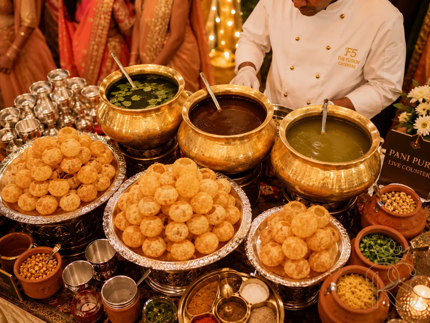 Indian wedding pani puri counter with silver thali trays, brass urns of flavoured pani, and a uniformed chef serving golgappas to guests