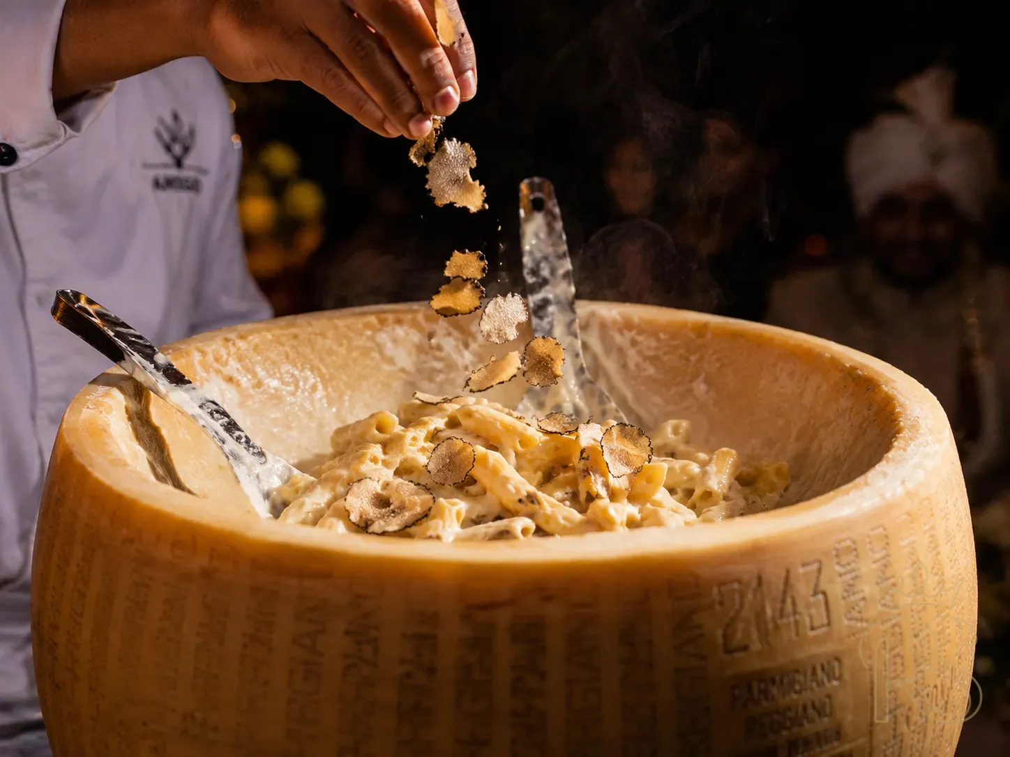 Macro close-up of a parmesan wheel pasta at an Indian wedding — hot pasta being tossed inside a scooped-out parmigiano reggiano wheel, cheese visibly melting into the pasta, steam rising, small white truffle shavings being added from above by a chef's hand, dramatic dark background, food photography, warm amber light, shallow depth of field