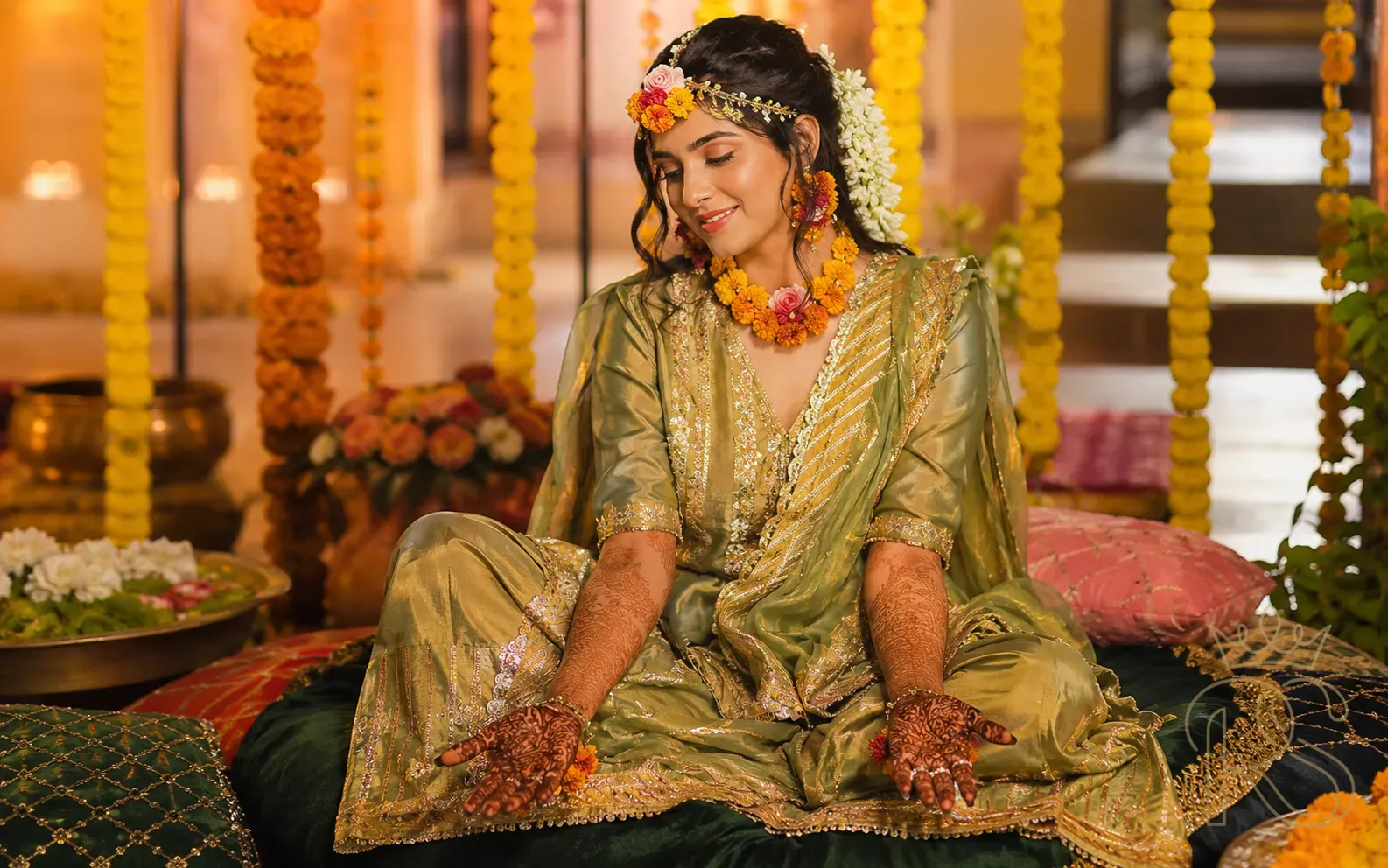 Indian bride at Mehendi ceremony in green and gold sharara with gota patti work, fresh mehndi on hands, flower jewellery and jasmine hair