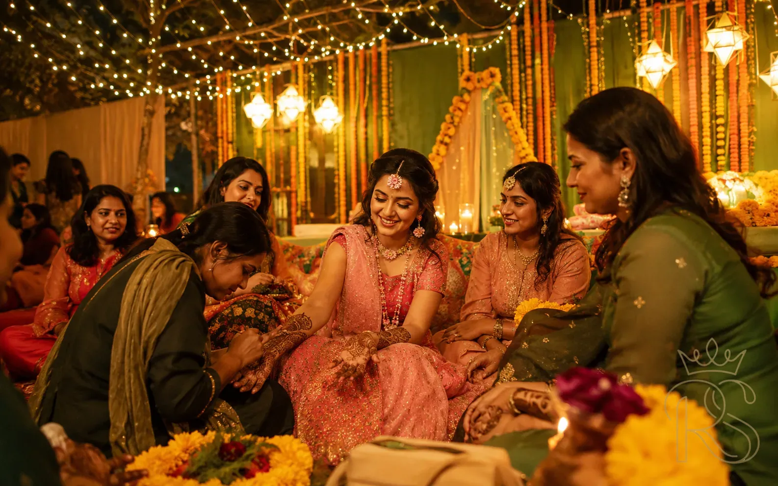 Indian Mehendi ceremony in the evening with bride in pink surrounded by guests, artist working on hand, string lights and floral décor