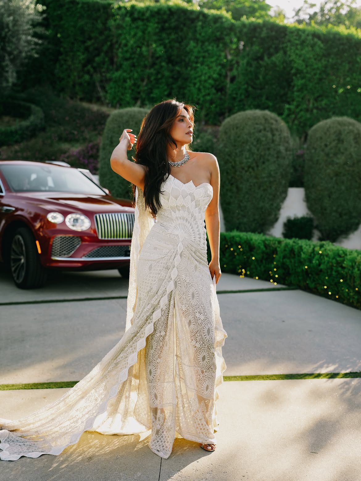 Priyanka Chopra in a strapless embroidered lace gown with a long train, standing beside a red car