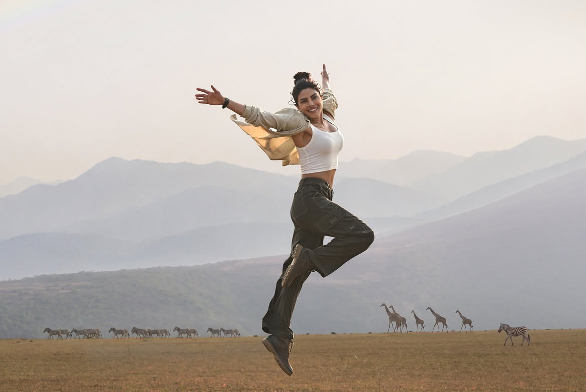 Priyanka Chopra jumping mid-air in a white tank top and cargo pants with zebras and giraffes in the background