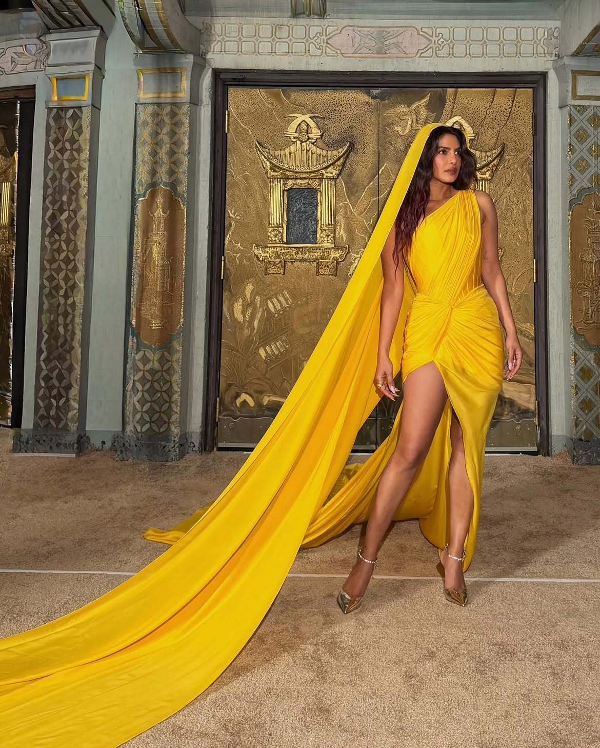 Priyanka Chopra in a bold yellow one-shoulder gown with a dramatic train, styled with a matching head-covering, in front of ornate gold doors
