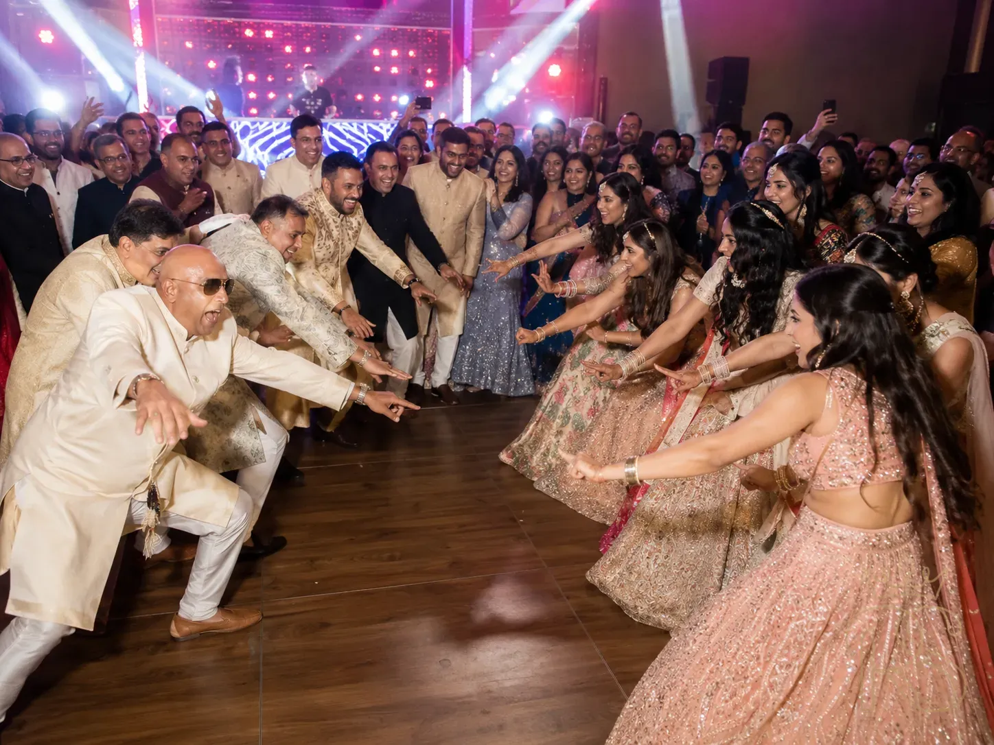 Indian wedding family dance battle at sangeet — a group of uncles in sherwanis on one side of the dance floor doing an exaggerated dance move, bridesmaids in lehengas on the opposite side countering with their own move, guests in a circle around them cheering, bright dance floor lighting, DJ booth visible in background, candid wedding photography, spontaneous joy, natural expressions of laughter and excitement