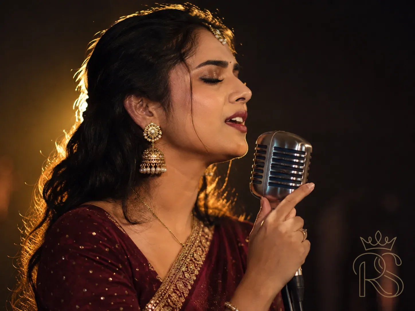 Close-up of a female Indian wedding singer performing at a sangeet — her face in profile against a dark background, holding a vintage microphone, eyes closed in emotional delivery, she wears a deep burgundy saree, stage lighting creating warm halo effect around her, shallow depth of field, concert photography style, intimate wedding performance moment