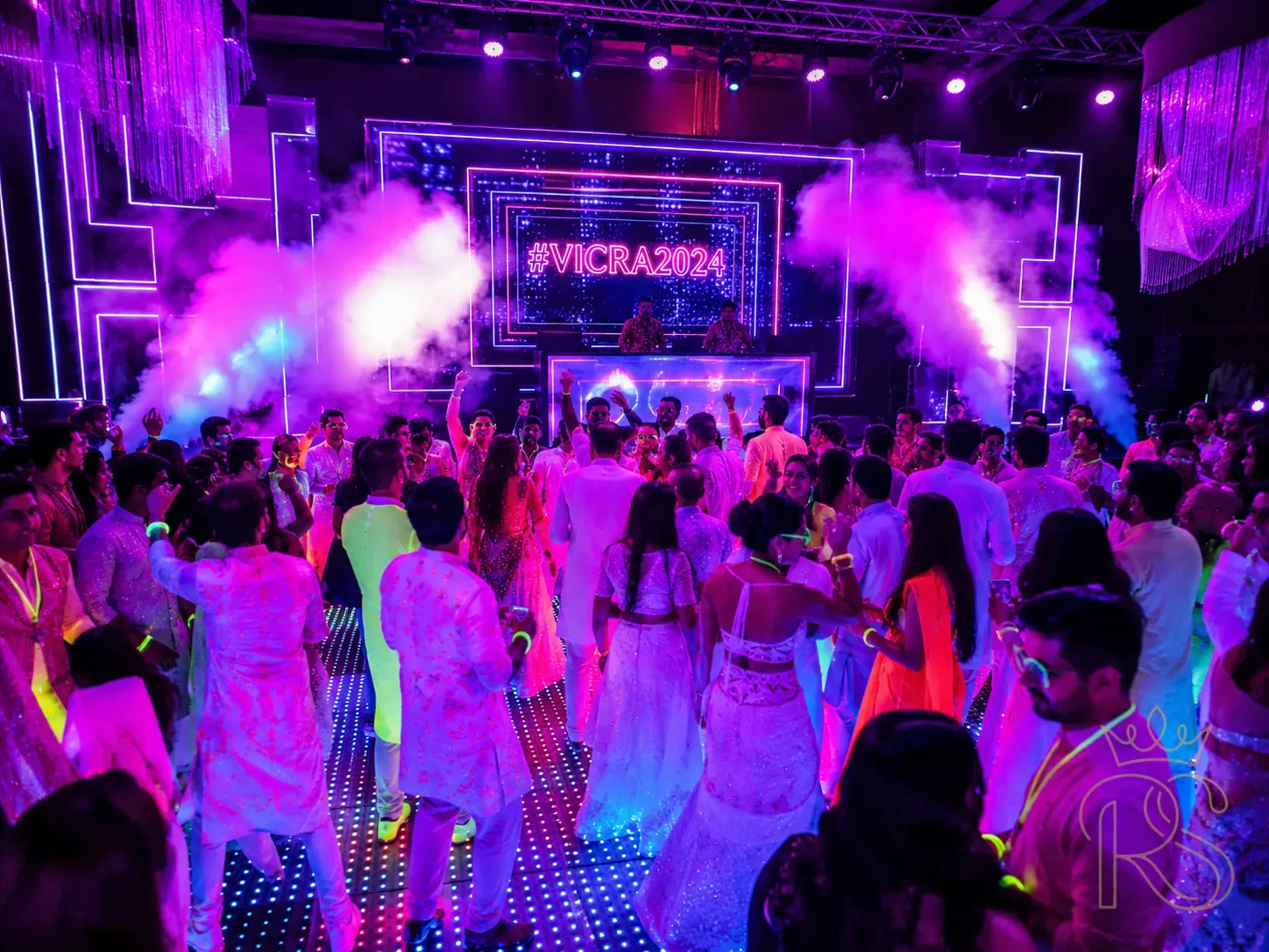 Indian wedding sangeet neon night theme — dark venue with vivid neon pink and purple LED lighting, guests in white and neon outfits glowing under UV light, a DJ booth with LED panels, dance floor with light-up tiles, fog machine effects, high energy wedding party atmosphere, wide-angle photography, electric colours, editorial style