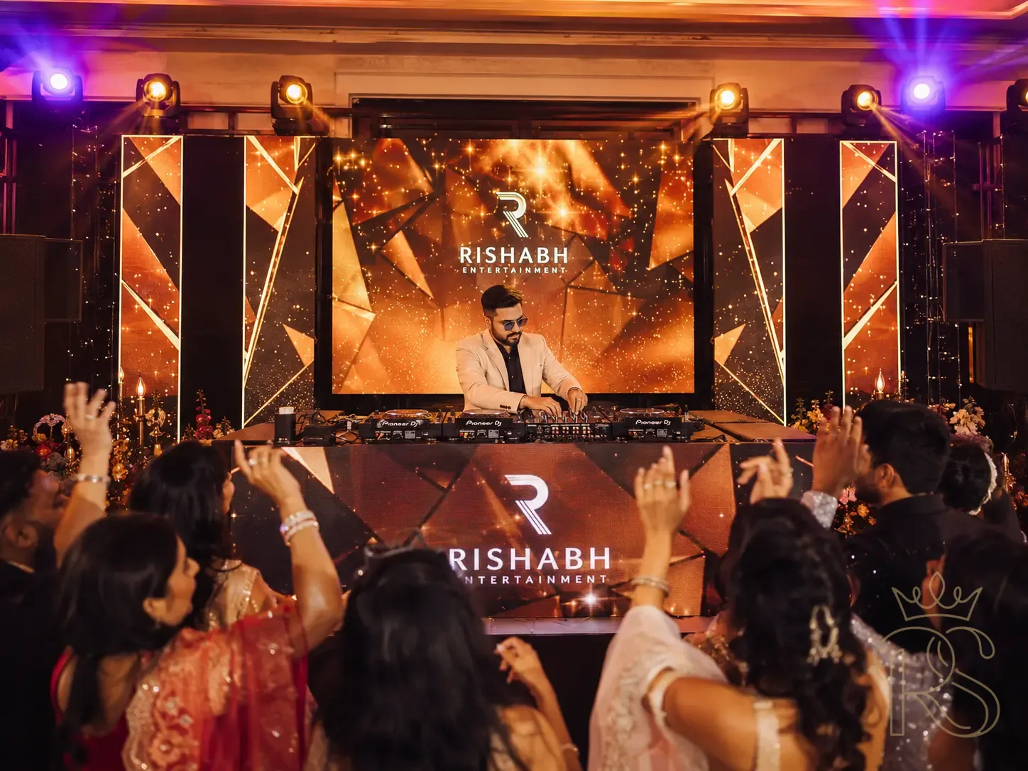 Professional DJ setup at an Indian wedding sangeet — a sleek DJ booth with dual CDJ decks and mixer, branded LED panels behind displaying abstract gold and red visuals, the DJ in a smart casual outfit adjusting the mix, warm amber and violet wash lighting, guests visible dancing in the foreground with backs to camera, professional wedding entertainment photography, wide angle, vibrant and energetic atmosphere