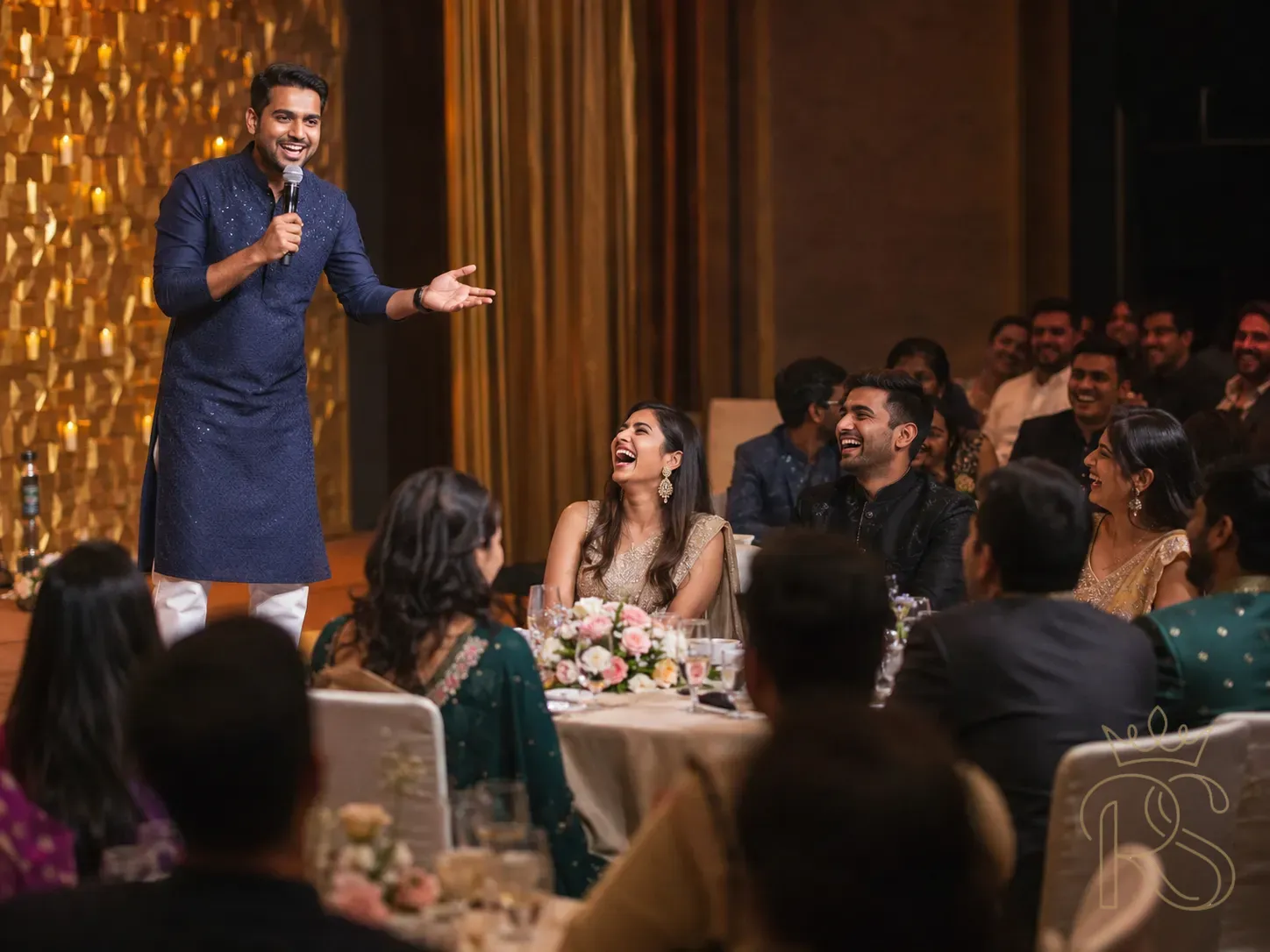 Indian wedding sangeet stand-up comedian performing on stage — a male comedian in a smart kurta holding a microphone, mid-gesture, guests at round tables in the foreground laughing openly, candid audience reaction shot, warm amber stage lighting, intimate wedding venue, professional wedding entertainment, photojournalism style, natural expressions, laughter visible on multiple guest faces
