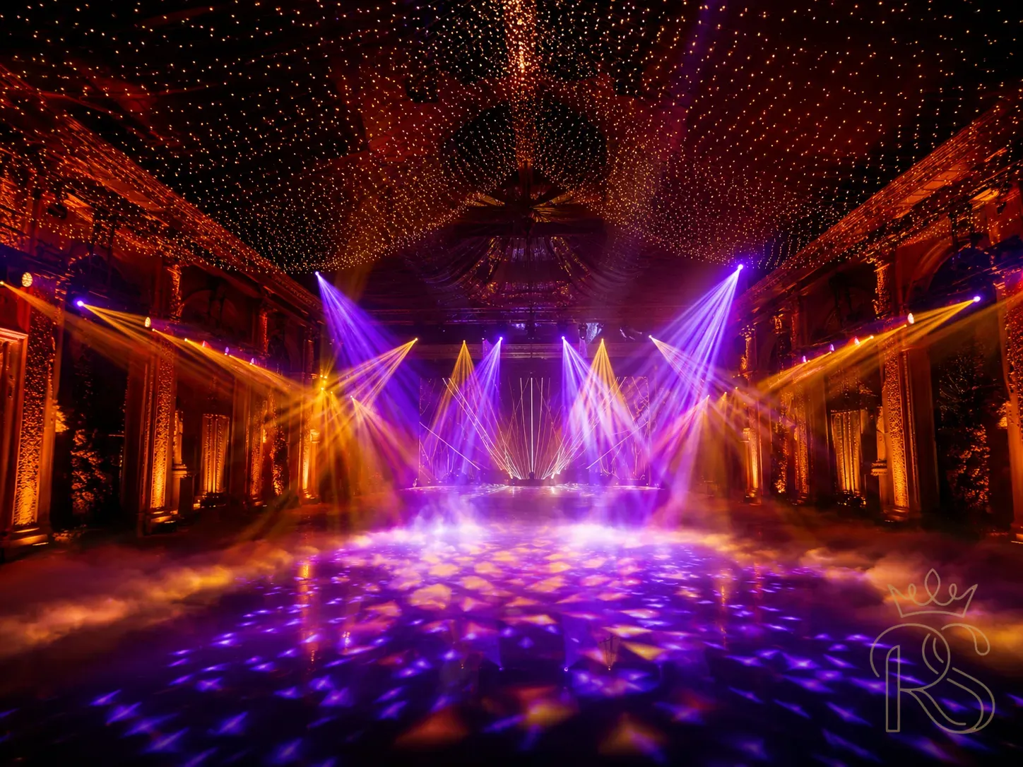 Breathtaking sangeet night lighting design — a wedding venue transformed by professional event lighting: warm amber uplights washing the walls, deep violet and gold spotlights crossing over the dance floor, a ceiling of white fairy lights forming a glowing canopy, laser effects at the far wall, theatrical fog at floor level, no guests visible — the empty venue showing the pure lighting design at its maximum, architectural photography, moody and electric, cinematic quality