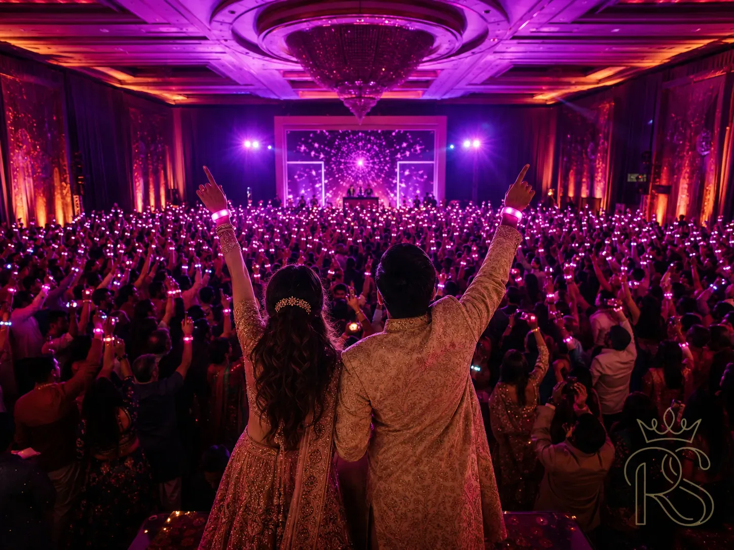 Indian wedding sangeet 2026 trend highlight — bride and groom with LED wristbands on their wrists that glow in sync with the music, a sea of guests all holding glowing wristbands creating a light show effect in the dark venue, DJ booth visible at the far end, wide angle shot showing the full crowd from the back of the venue, immersive concert-like atmosphere, warm magenta and purple glowing tones, editorial wedding photography
