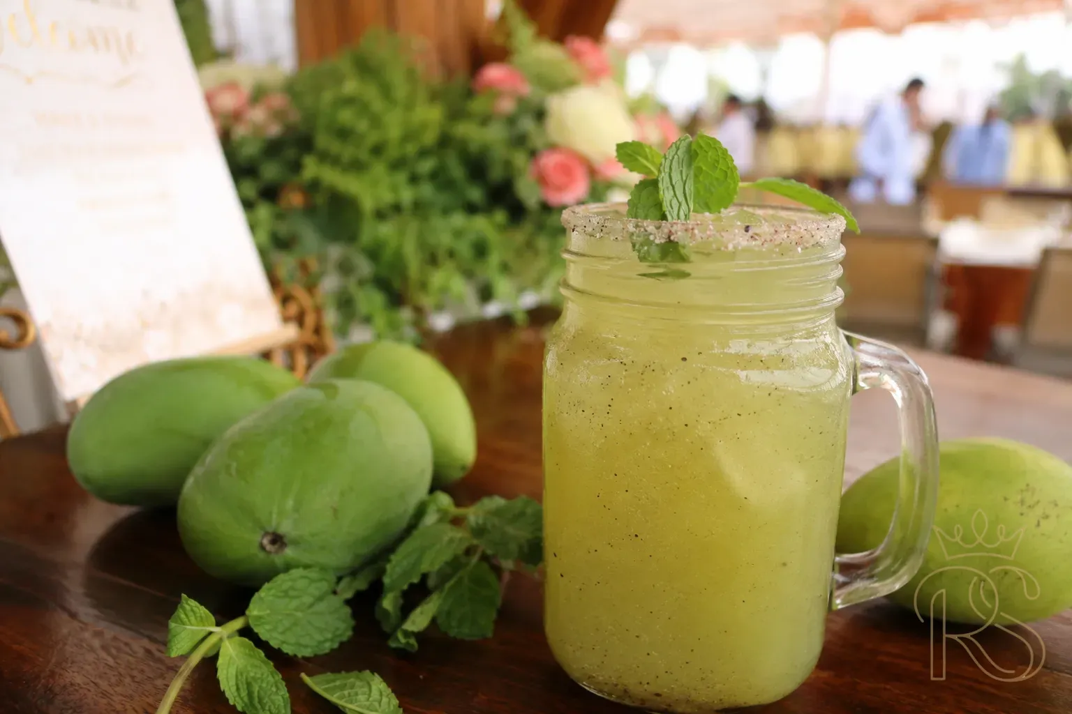 Vibrant green raw mango panna cooler in a mason jar garnished with mint leaves and black salt rim, surrounded by raw mangoes on a wooden bar surface at a summer wedding