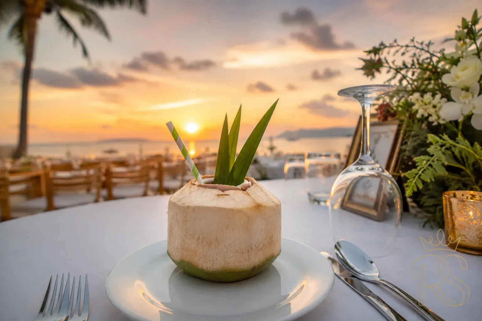 Elegant coconut and pandan cooler served in a halved coconut shell with a paper straw and pandan leaf garnish, placed on a draped white wedding reception table against a beachside sunset backdrop