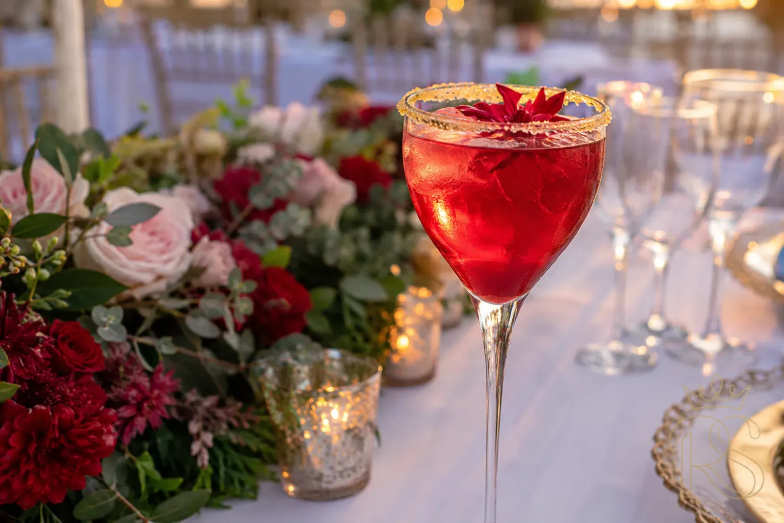 Deep ruby red hibiscus and ginger sparkling cooler in a crystal wine glass with a candied ginger rim, fresh hibiscus flower floating in the drink, wedding reception table setting in the background