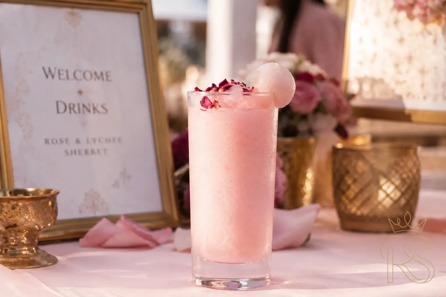 Tall glass of rose and lychee sherbet garnished with dried rose petals and a lychee on the rim, served at an elegant Indian wedding welcome station, soft pink tones